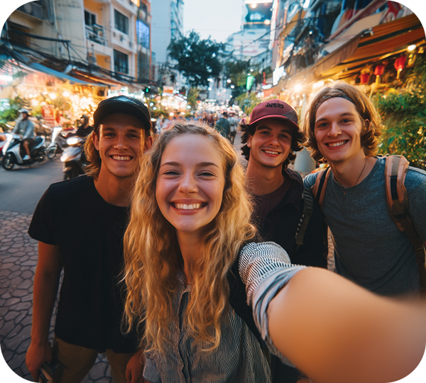 Four young adults smile for a selfie on a lively, bustling city street lined with market stalls and bright lights in the evening.