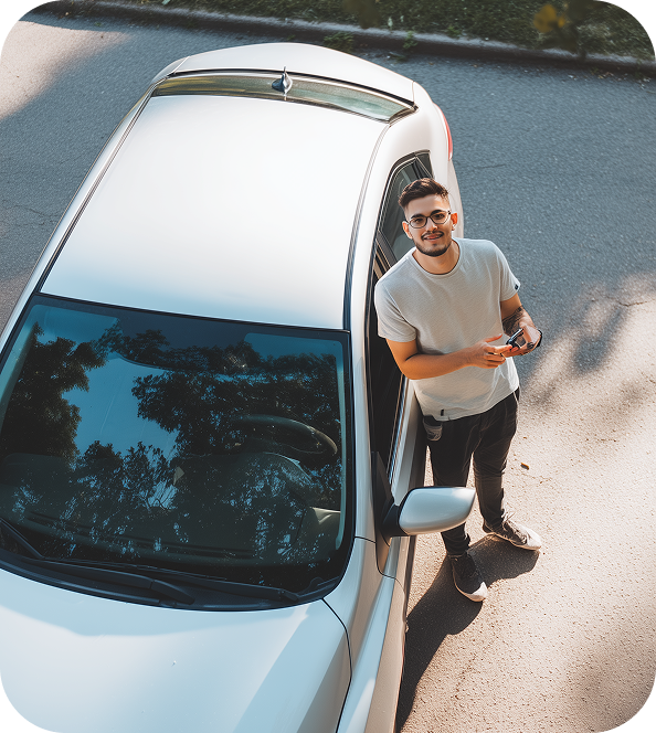 Man standing next to a white car on a paved road, holding car keys and looking up toward the camera.